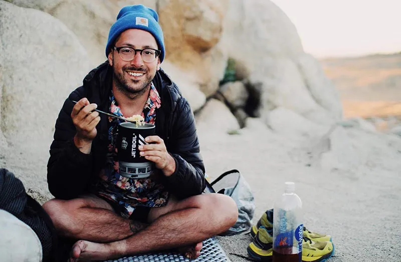 A hiker sits cross-legged, spooning food out of a Jetboil cook pot and smiling for the camera. 