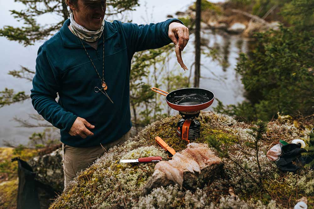 Man adding fish to a sizzling Jetboil summit skillet with TrailWare utensils while outside
