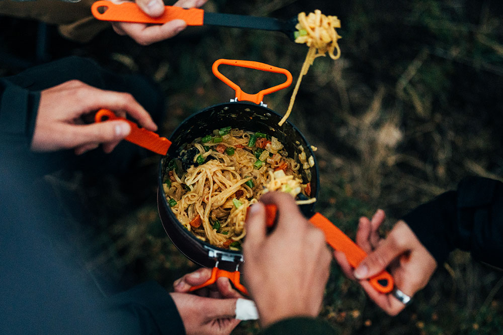 Group of hands using Jetboil TrailWare utensils reaching for delicious cooked mean inside Jetboil TrailCook 2.0L cooking system