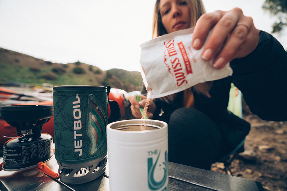 Close up shot of a person pouring Swiss Miss instant cocoa mix into a cup of camp coffee with a Jetboil Flash in the background.