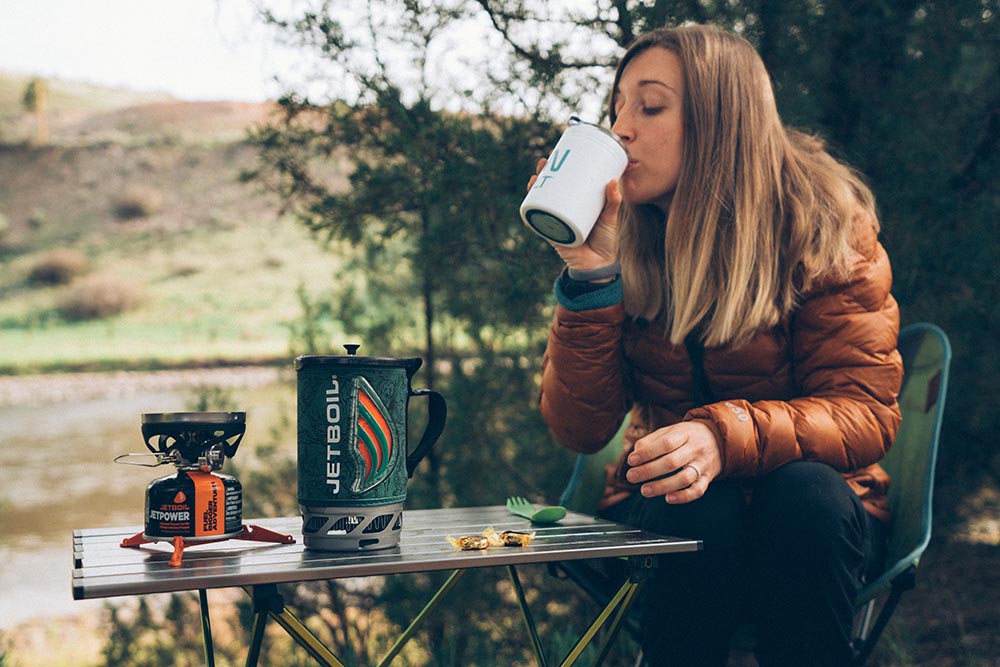 A woman takes a sip from a camp coffee while sitting next to a camp table with a Jetboil Flash system next to her on the table. 