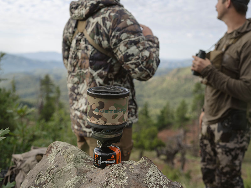 A camo Jetboil MiniMo with two hunters glassing in the background.