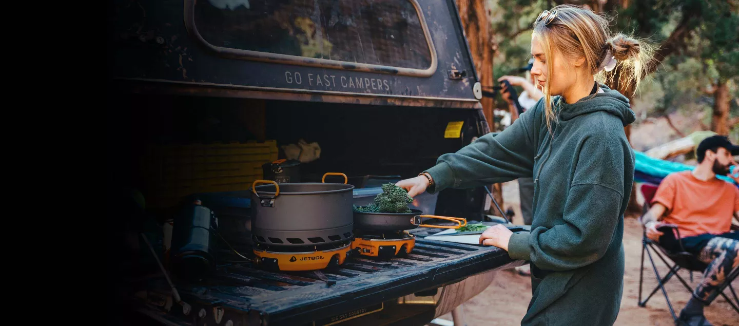 A woman cooks on a two burner camp stove on the tailgate of a truck.