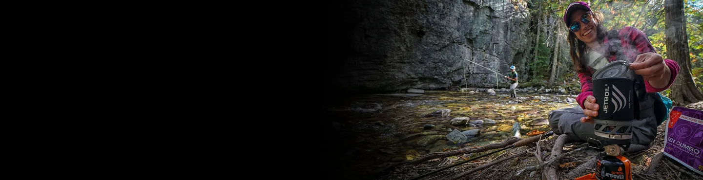 Woman lifting the lid off a Jetboil MicroMo cooking system with seam coming out sitting near a riverbend