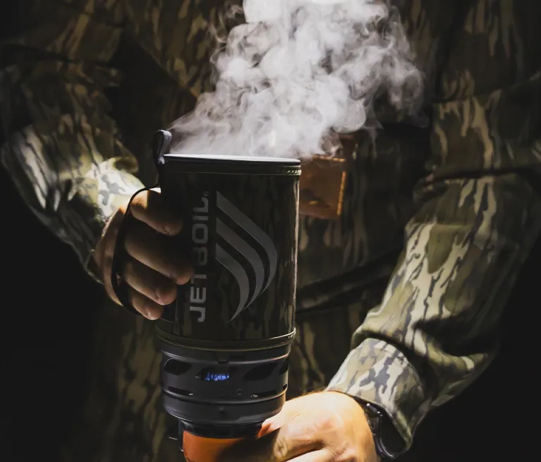 Holding a steaming Jetboil Flash 1.0L fast boil system in mossy oak bottomland